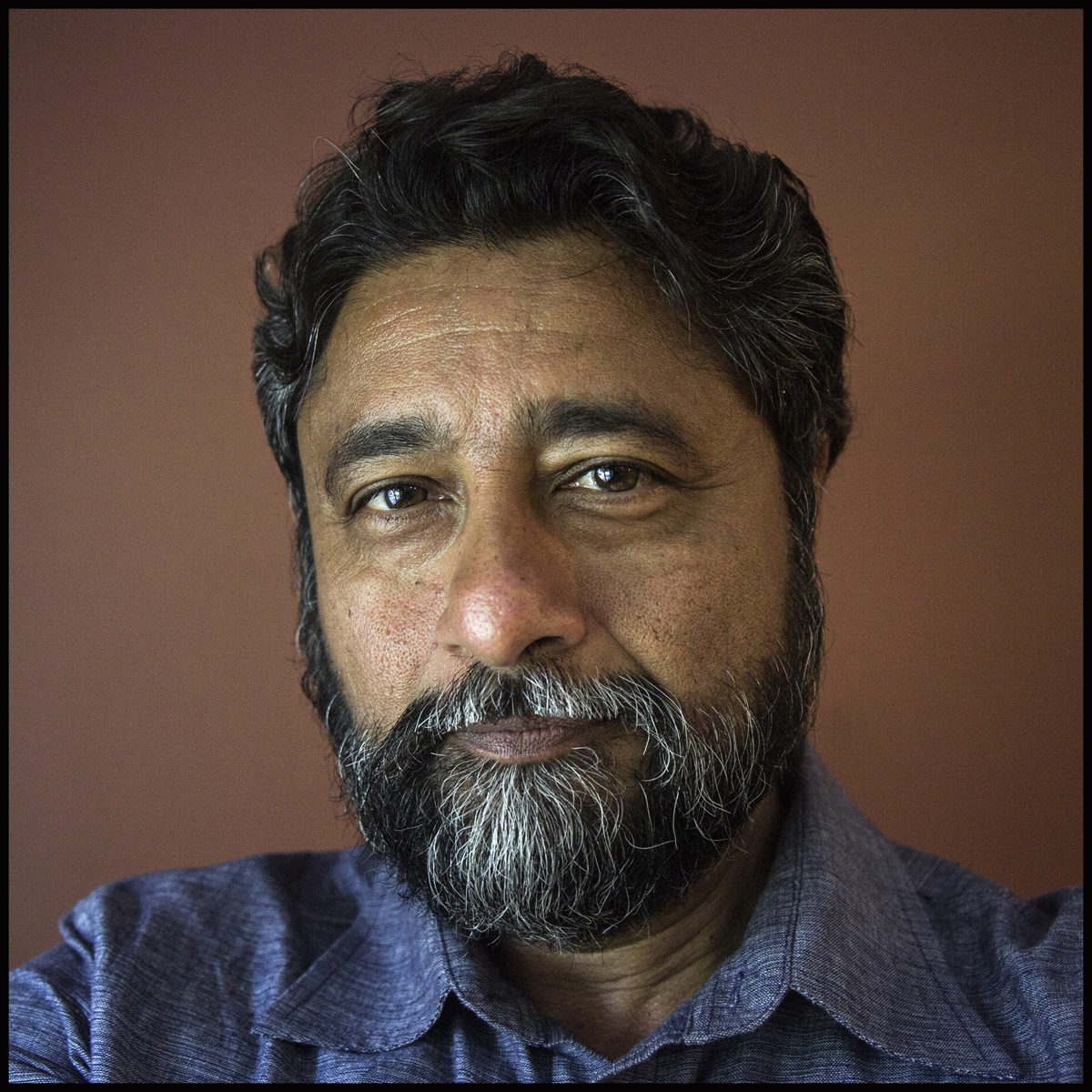 Headshot of a person with dark hair and a gray beard, wearing a collared shirt, against a plain background.