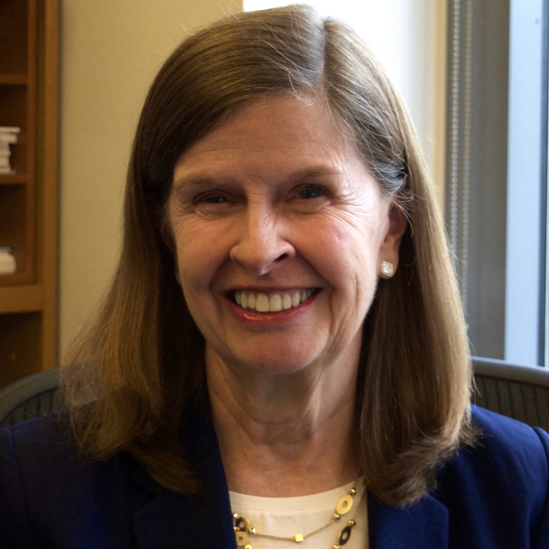 Headshot of a smiling person with shoulder-length hair, wearing a blue jacket and a gold necklace.