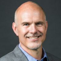 Headshot of a smiling individual with a bald head, light facial hair, and wearing a gray suit jacket.