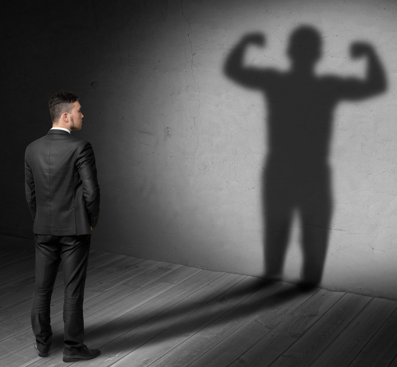 A person in a suit is standing in a room, gazing at a wall where their shadow appears muscular and flexed. The image represents self-perception and inner strength.