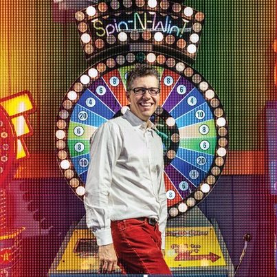 A person smiling in front of a colorful "Spin-N-Win" wheel, with vibrant lights and a game show atmosphere.