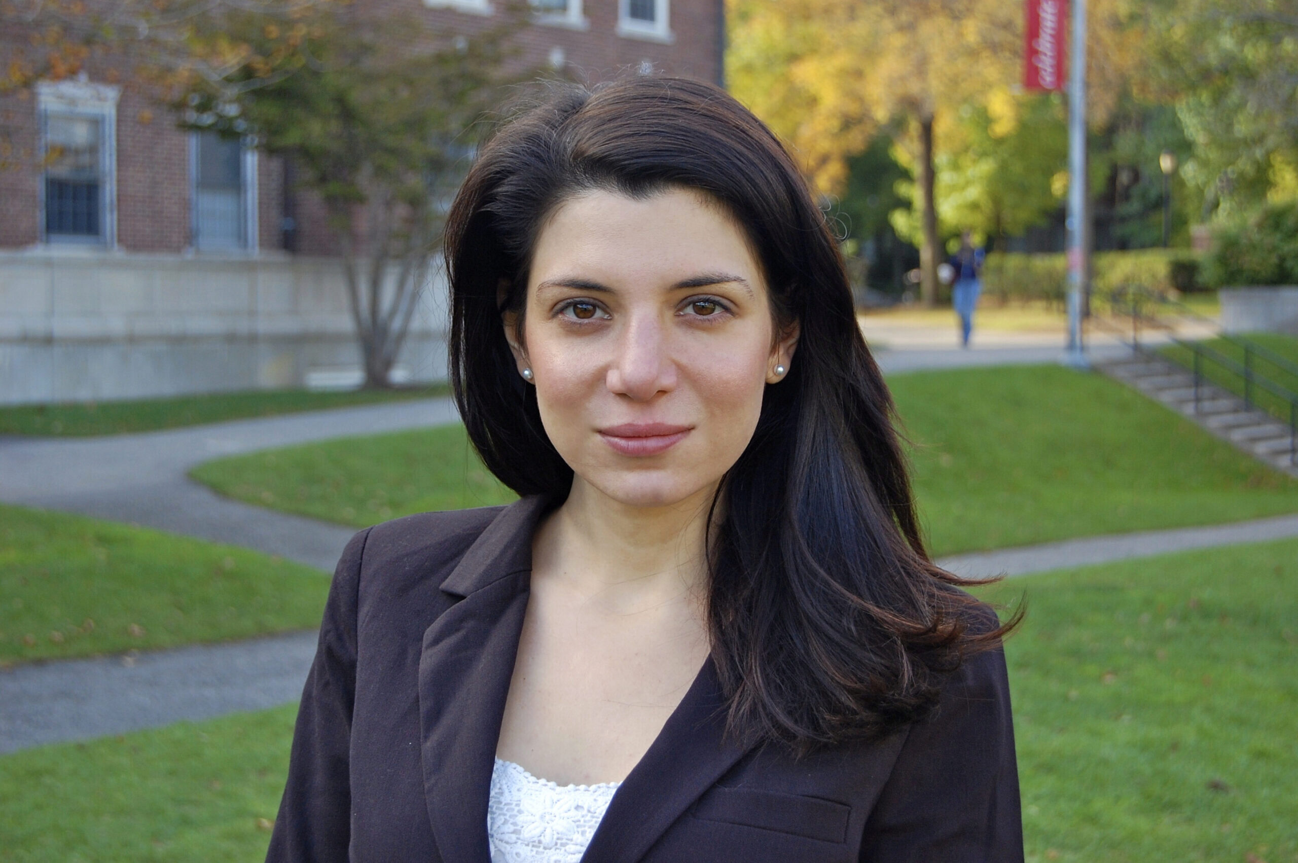 Headshot of a person with long dark hair standing outdoors, wearing a brown blazer. The background shows a grassy area with trees and a building.