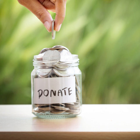 A hand dropping a coin into a jar labeled "Donate," filled with other coins, against a blurred green background.