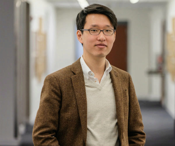 A headshot of a person wearing glasses, a brown blazer, and a white shirt, standing in a hallway.