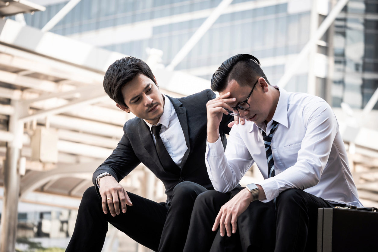 A person in a suit is sitting next to another who appears distressed, offering comfort by placing a hand on their shoulder.