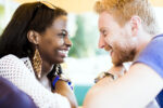 A close-up of a smiling couple facing each other with an affectionate gaze. Both wear watches, and one wears large earrings.