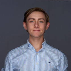 Headshot of a person with short brown hair wearing a blue and white striped button-up shirt. The background is a plain dark gray.