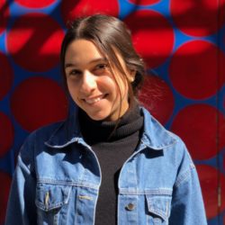 A person smiling in a headshot, wearing a denim jacket and black turtleneck, standing in front of a red and blue circular patterned background.