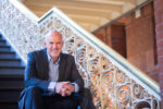 A person sitting on a stairway, wearing a suit and smiling. The staircase features an ornate, decorative railing, and the background includes red brick walls and architectural details.