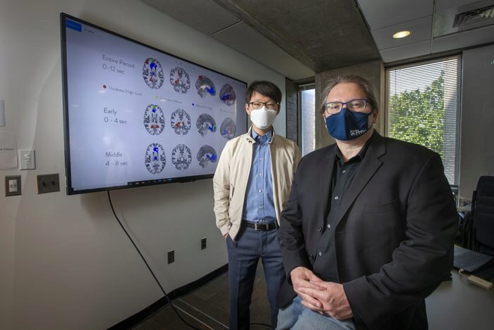 Two individuals wearing masks stand in an office near a large screen displaying brain images and data, suggesting a research or scientific setting.