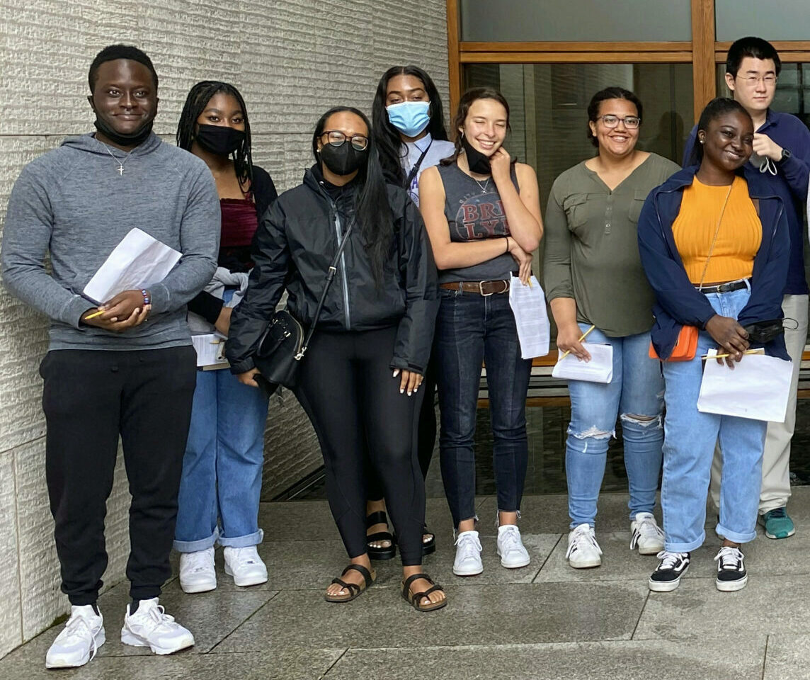 A group of people standing together, some holding papers. Most are smiling, and some are wearing face masks. They are dressed in casual attire.