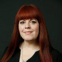 This image is a headshot of a person with long, red hair and fringe, wearing a black top and a silver necklace, set against a dark background.