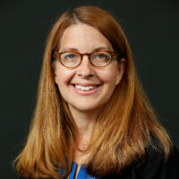 A headshot of a person with long brown hair and glasses, smiling, set against a dark background.