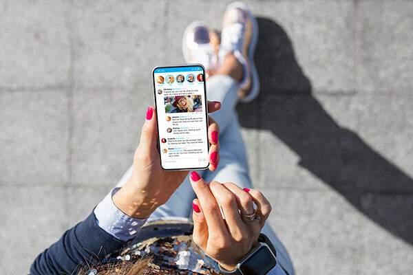 A person holding a smartphone displaying a social media app with several messages. They are sitting outdoors, wearing casual clothes and sneakers.
