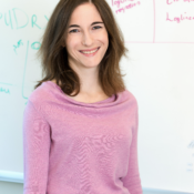 A person smiling in front of a whiteboard with writing and diagrams. The individual is wearing a pink sweater.