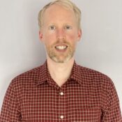 Headshot of a person with short light hair, wearing a red plaid shirt, against a plain background.