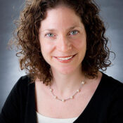 A headshot of a person with curly hair, smiling, wearing a black top and a pearl necklace against a neutral background.