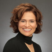 A headshot of a smiling person with curly brown hair wearing a dark high-collared top against a neutral background.