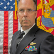 A person in a military uniform with medals, standing in front of an American flag and a Marine Corps flag.