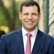 Headshot of a smiling person in a blue suit and pink tie, standing outdoors in front of a brick wall with blurred greenery in the background.