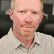 Headshot of a person with short blond hair and a beard, wearing a light gray collared shirt, set against a patterned background.