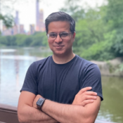 A person wearing glasses and a smartwatch stands with arms crossed in front of a lake with a city skyline and greenery in the background.