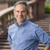 A person in a blue shirt stands outdoors with hands on hips, in front of a blurred background featuring a staircase and greenery.