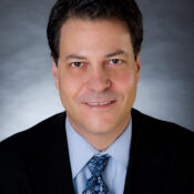 A headshot of a person wearing a black suit, light blue shirt, and a patterned blue tie, set against a gray background.