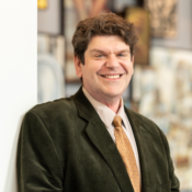 A headshot of a person smiling, wearing a suit jacket and tie, standing in front of an abstract, blurred background.