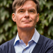 A headshot of a person with short hair, wearing a blue button-up shirt and a dark sweater, set against a leafy green background.