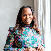 A person smiling while wearing a colorful, floral-patterned dress, standing near a window with white curtains.