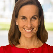 A headshot of a smiling person with brown hair, wearing a red top, a necklace, and small earrings. The background is softly blurred.