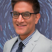 A headshot of a person wearing glasses and a light gray suit over a checkered shirt with a blue tie, set against a colorful, swirling background.