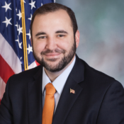 Headshot of a person in a suit and orange tie, smiling, in front of a U.S. flag.