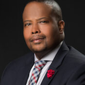 A professional headshot of a person wearing a dark suit, patterned tie, and red lapel pin, against a dark background.