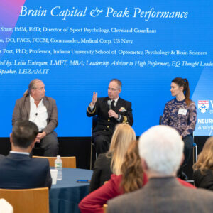 A panel discussion on "Brain Capital & Peak Performance" with three speakers seated on stage. The backdrop shows event details, and attendees are observed in the audience.