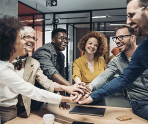 A diverse group of people in an office setting, smiling and stacking their hands together, representing teamwork and collaboration.