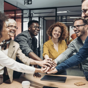 A diverse group of people in an office setting, smiling and stacking their hands together, representing teamwork and collaboration.