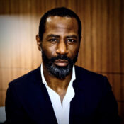 Headshot of a person with a beard, wearing a dark suit and white shirt, set against a wooden wall background.