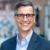 Headshot of a person wearing glasses, smiling, and dressed in a blue blazer and checkered shirt. The background is softly blurred, featuring some buildings.