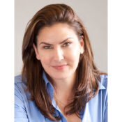 Headshot of a person with long brown hair wearing a blue shirt, smiling softly at the camera against a neutral background.