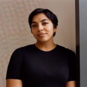 A person with short hair and a calm expression, wearing a black shirt, stands against a patterned wall.