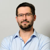 Headshot of a person with short dark hair, glasses, and a beard, wearing a light blue shirt, with arms crossed and a neutral expression.