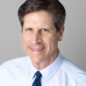 Headshot of a person with short dark hair, wearing a white shirt and blue tie, smiling gently against a neutral background.