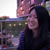 A person with long dark hair smiling near a balcony or patio area, with outdoor string lights and people dining in the background.