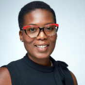 A headshot of a person wearing red and black glasses, with short hair and a black top, smiling at the camera against a light background.