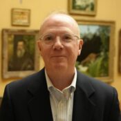 A headshot of a person wearing glasses and a suit jacket, standing in front of a wall adorned with framed paintings.