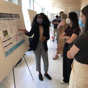 A person wearing a mask presents research information on a poster to a small group of individuals, who are also masked, in a bright indoor setting.