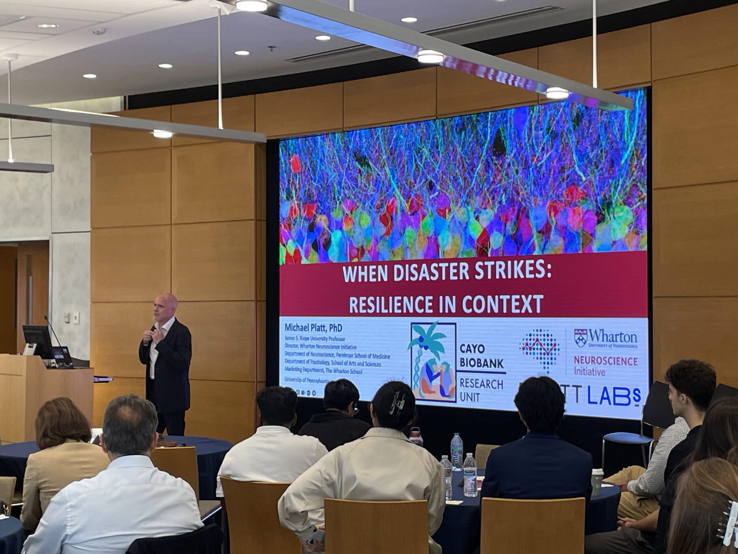 A speaker stands in front of an audience giving a presentation titled "When Disaster Strikes: Resilience in Context". The display screen features colorful neural imagery and logos from Wharton Neuroscience Initiative, Cayo Biobank, and related organizations. The setting