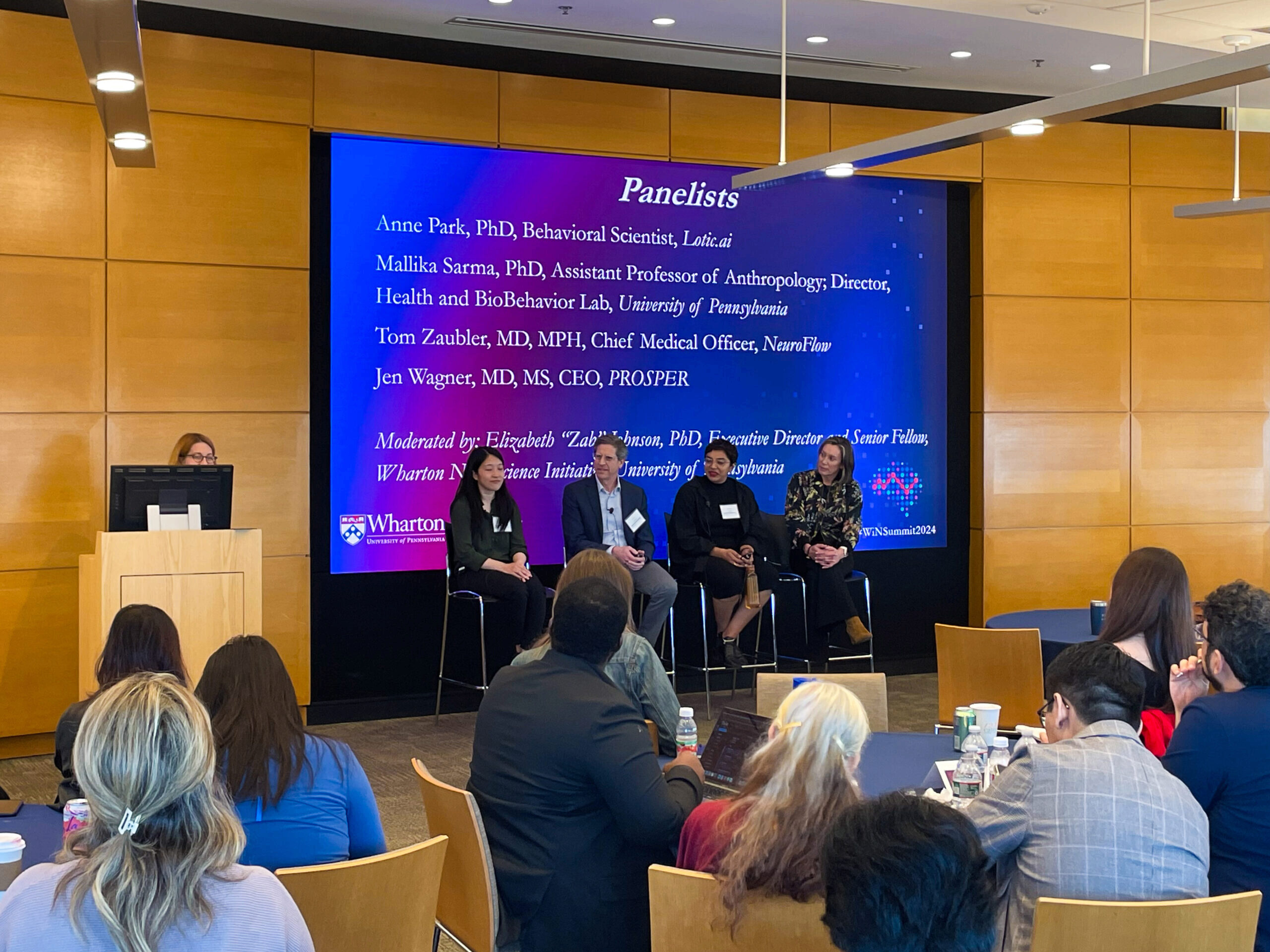 A panel discussion at an academic or professional event shows several panelists seated on stage with a moderator at a podium. A large screen behind them displays their names, titles, and affiliations. An audience is seated in front, attentively listening.